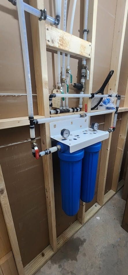 Wall-mounted plumbing manifold with white piping and two blue filter canisters in an unfinished wooden utility wall