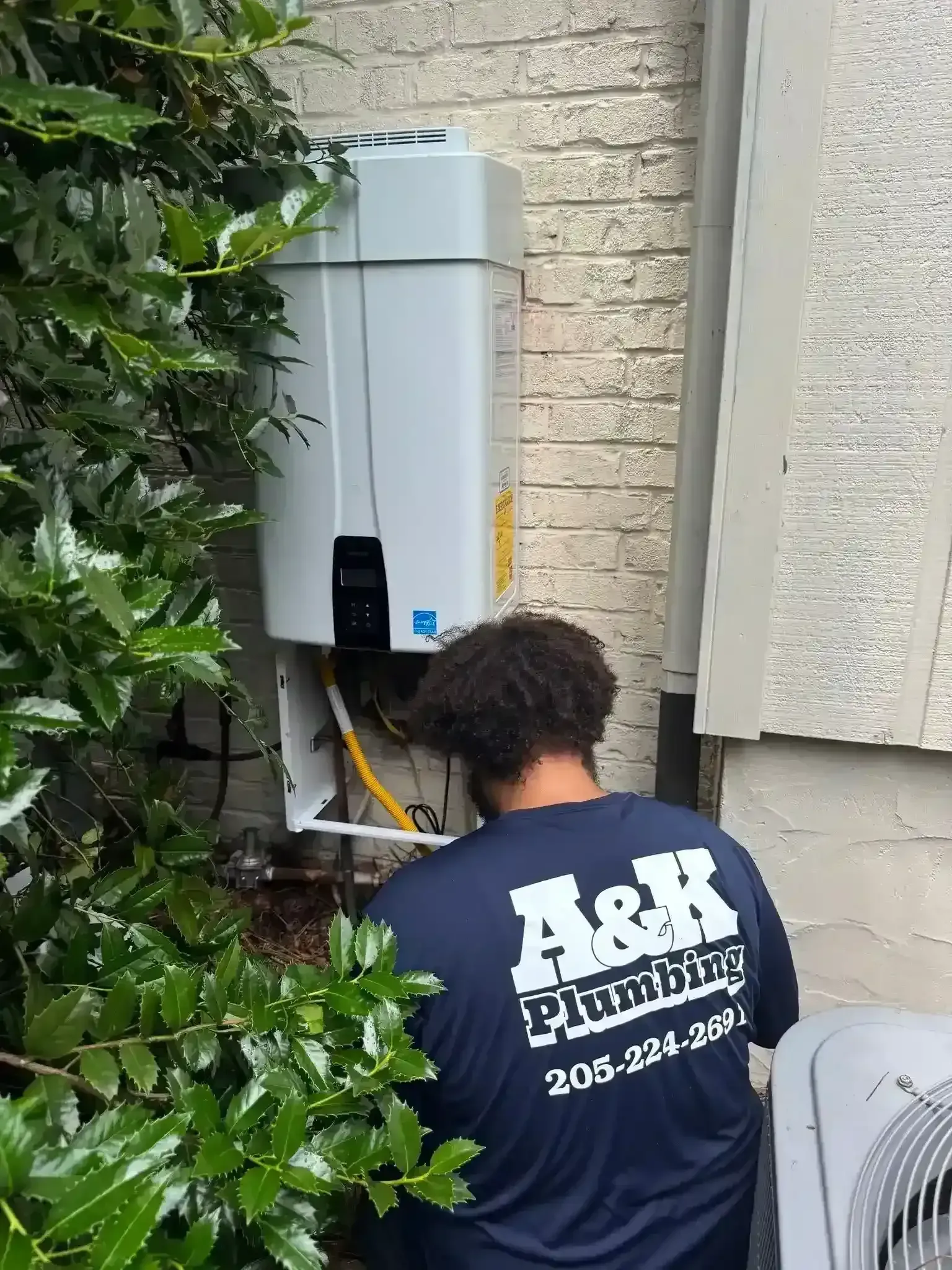Person working on an outdoor electrical box beside a brick wall and bushes