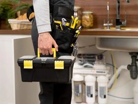 Person holding a black and yellow toolbox in a kitchen or utility area near a sink and water filters