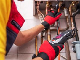Technician in red gloves inspecting plumbing under a sink with a digital meter.