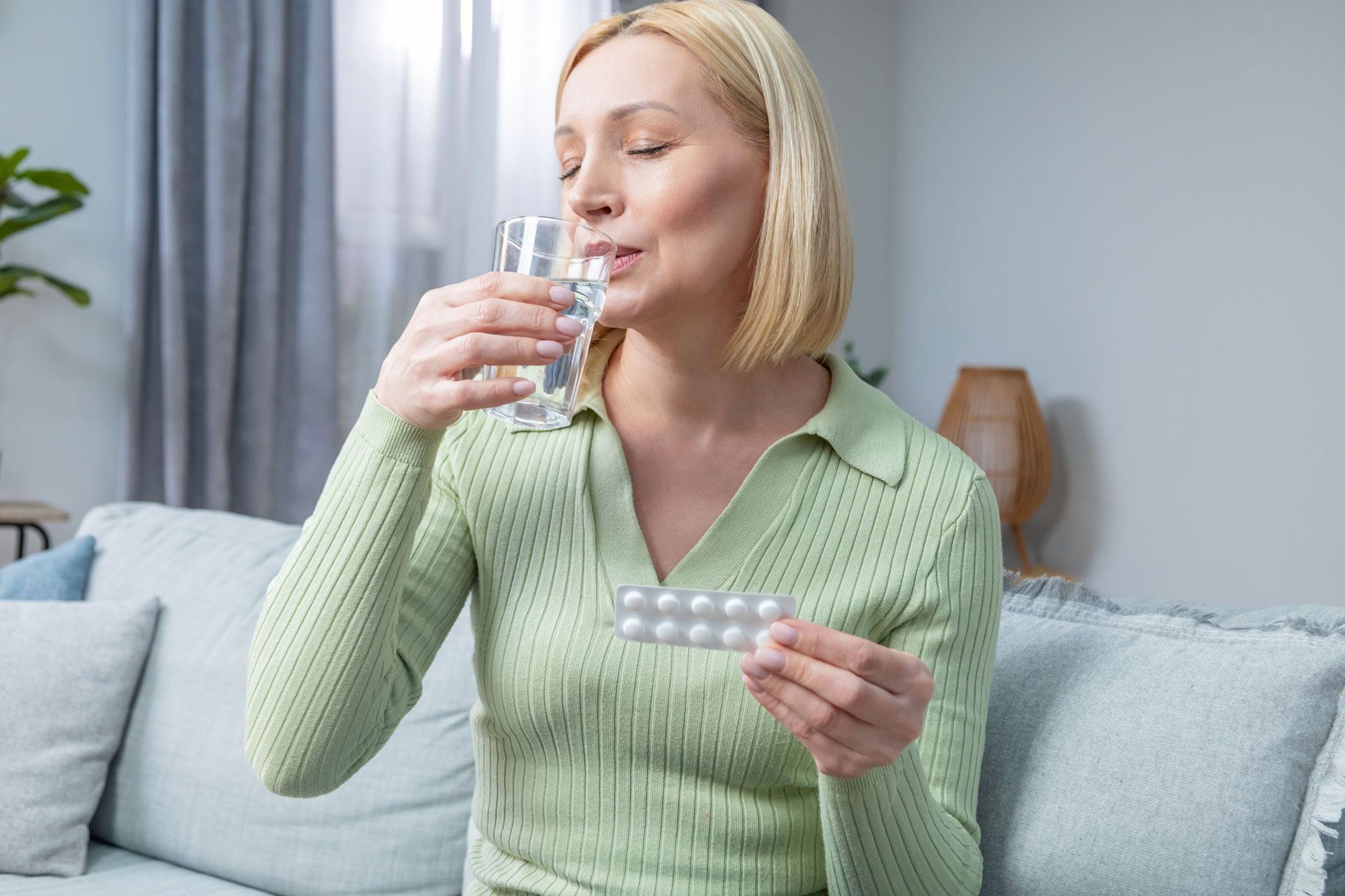 Woman sitting on a couch, drinking water and holding a blister pack of pills