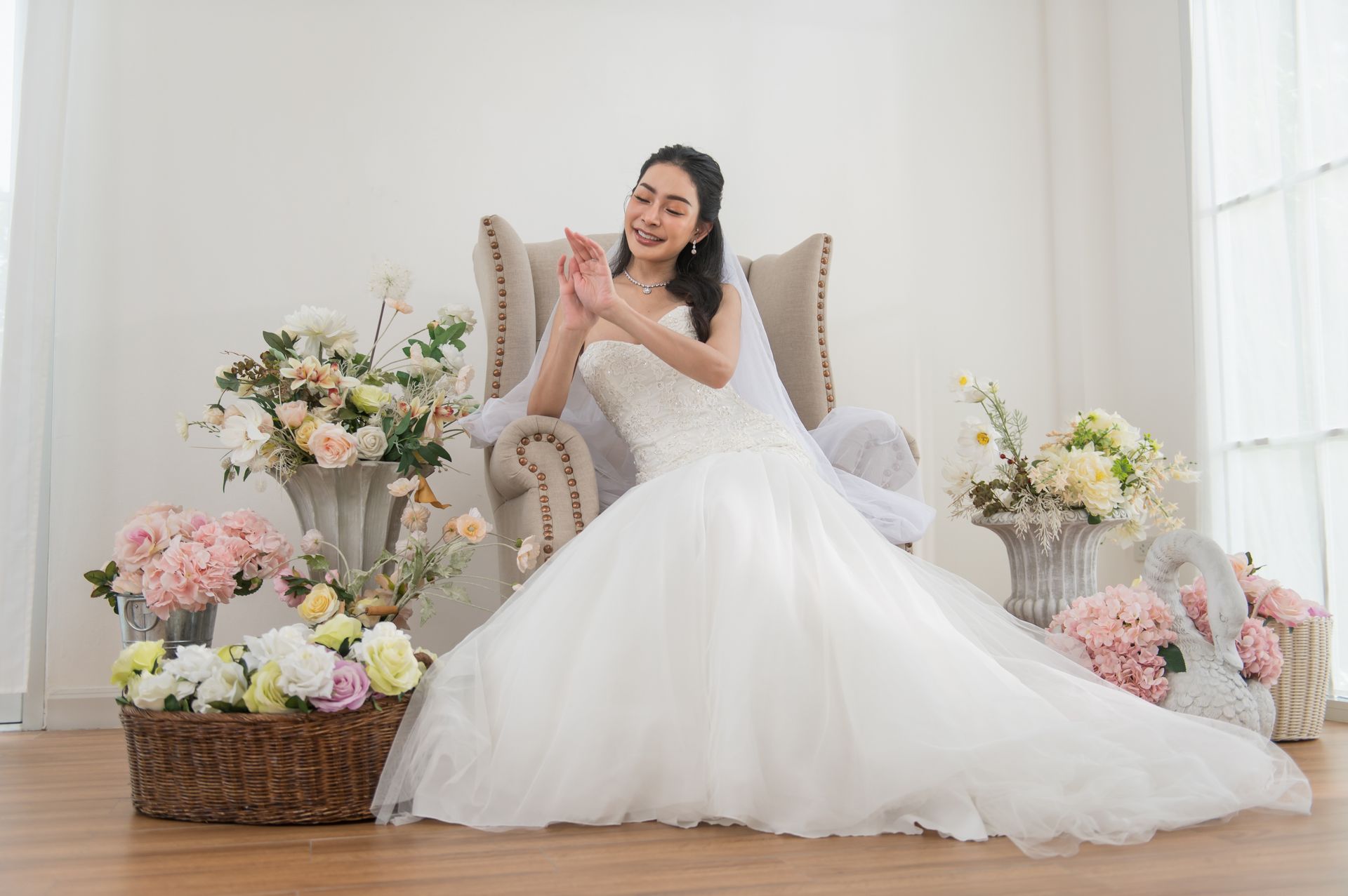 Bride in a white gown seated on a chair, surrounded by pastel floral arrangements in a bright room