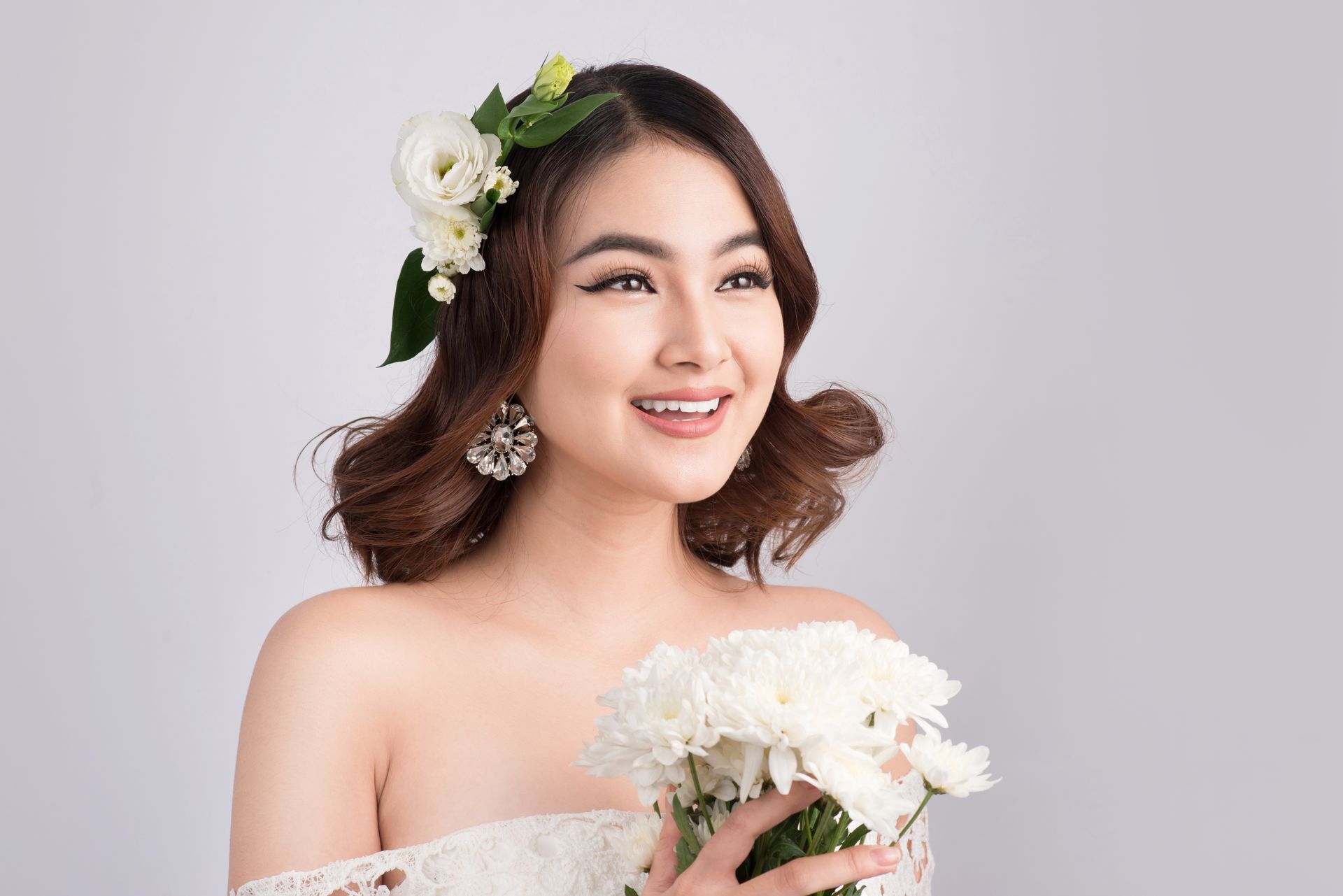 Smiling bride holding a white bouquet, wearing an off-shoulder gown and floral hair accessory.