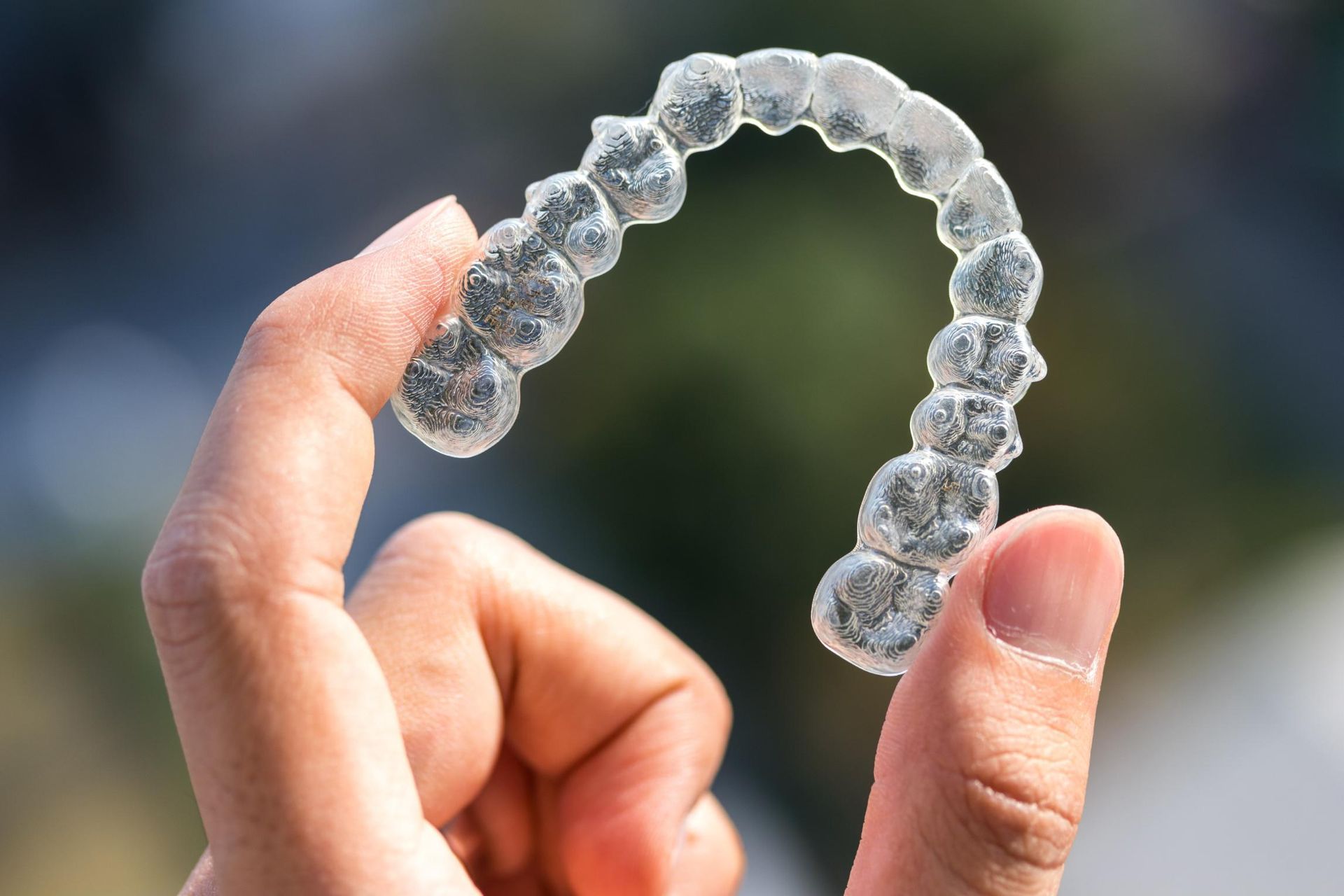 Hand holding a clear aligner tray in front of a blurred outdoor background