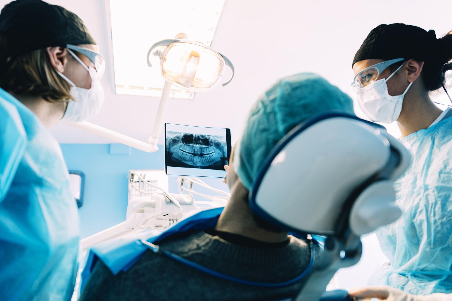 Surgeons in blue scrubs operate under bright lights, viewed from behind with a monitor showing the procedure
