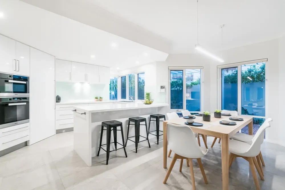 A bright, modern kitchen featuring white cabinets, a central island with three stools, and a wooden dining table nearby. — George Pappas Painting In Buderim, QLD