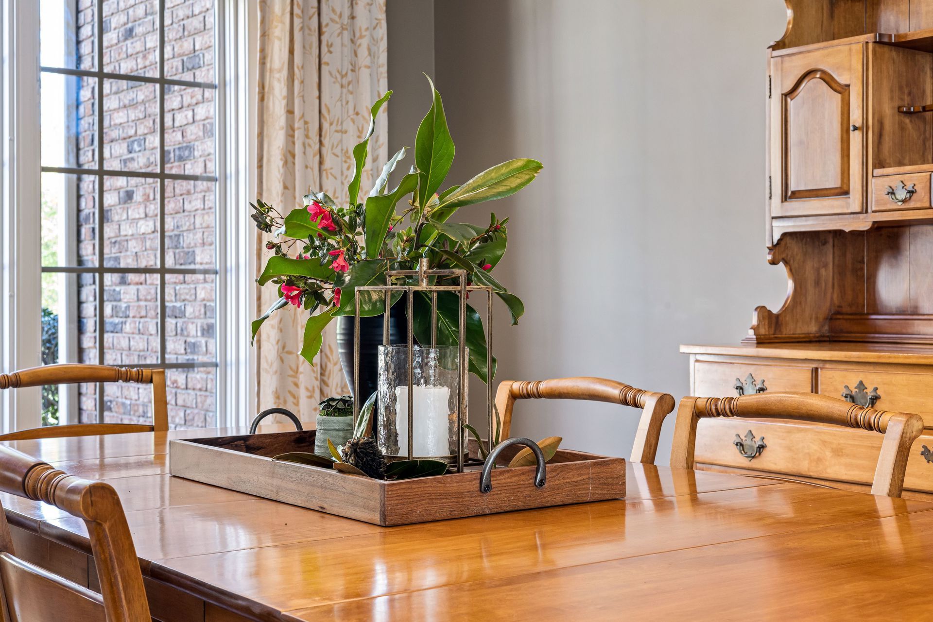 Dining room table with a potted plant centerpiece beside a sunlit window and wooden hutch.