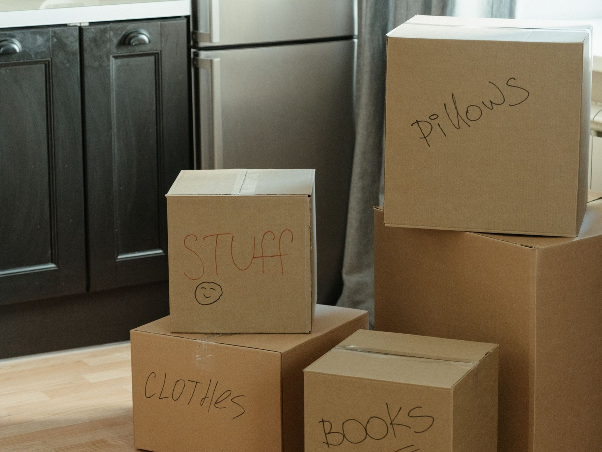 Cardboard moving boxes labeled pillows, books, clothes, and a lamp in a kitchen.