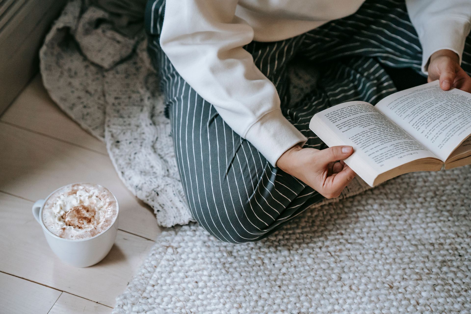 Person reading a book on a rug beside a cup of coffee and a gray blanket