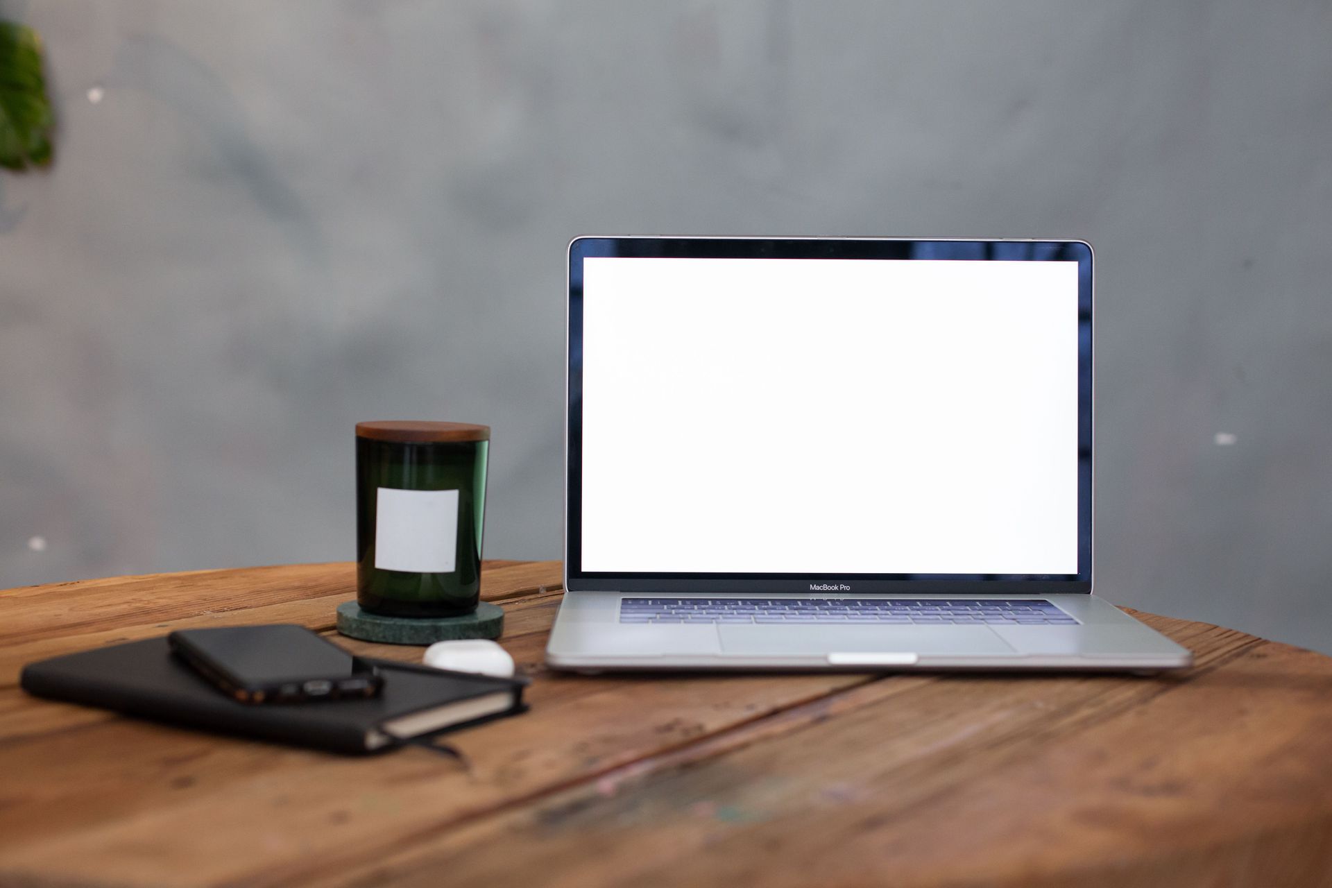 Laptop on a wooden desk with a phone, candle, and notebook against a gray wall.