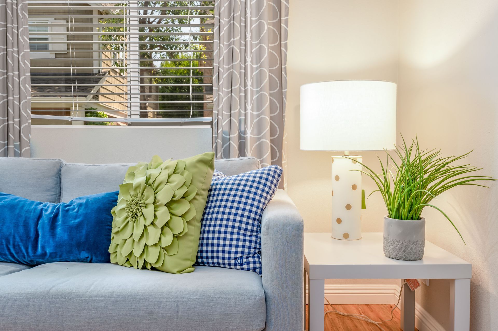 Living room corner with gray sofa, colorful pillows, side table, lamp, and plant by a window