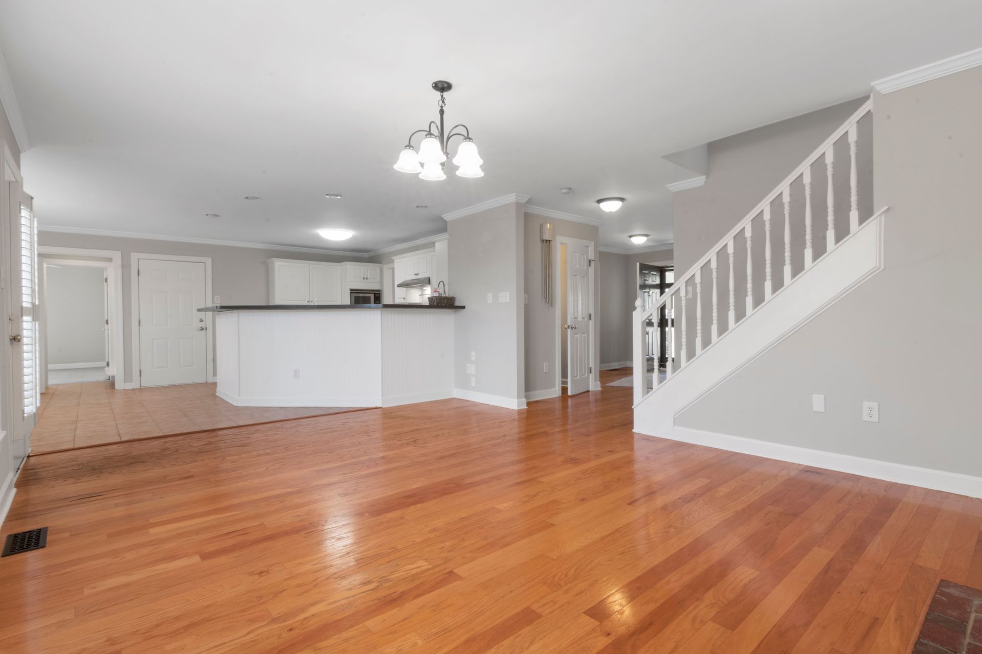 Empty living room with hardwood floors, white walls, staircase, and open kitchen area