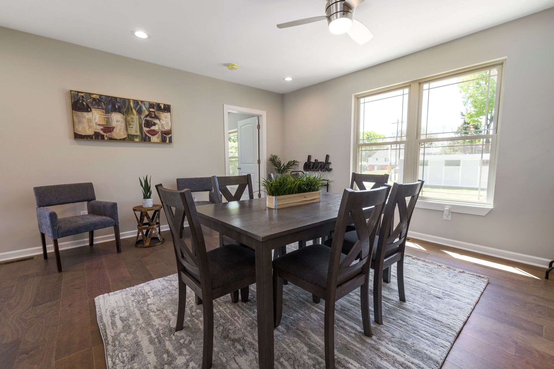 Dining room with a dark wood table, chairs, gray rug, and bright windows overlooking a backyard.