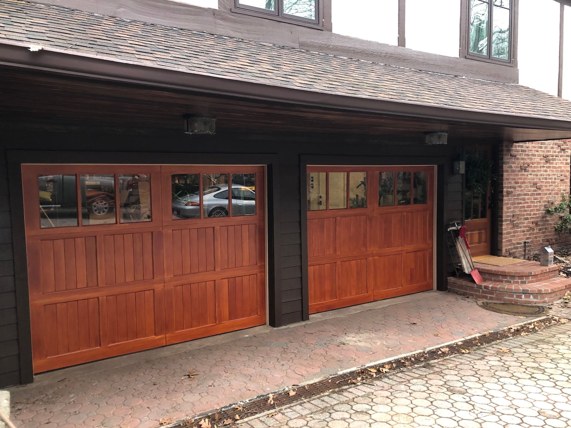 Two wooden garage doors under a shingled roof, with a brick wall and stone driveway.