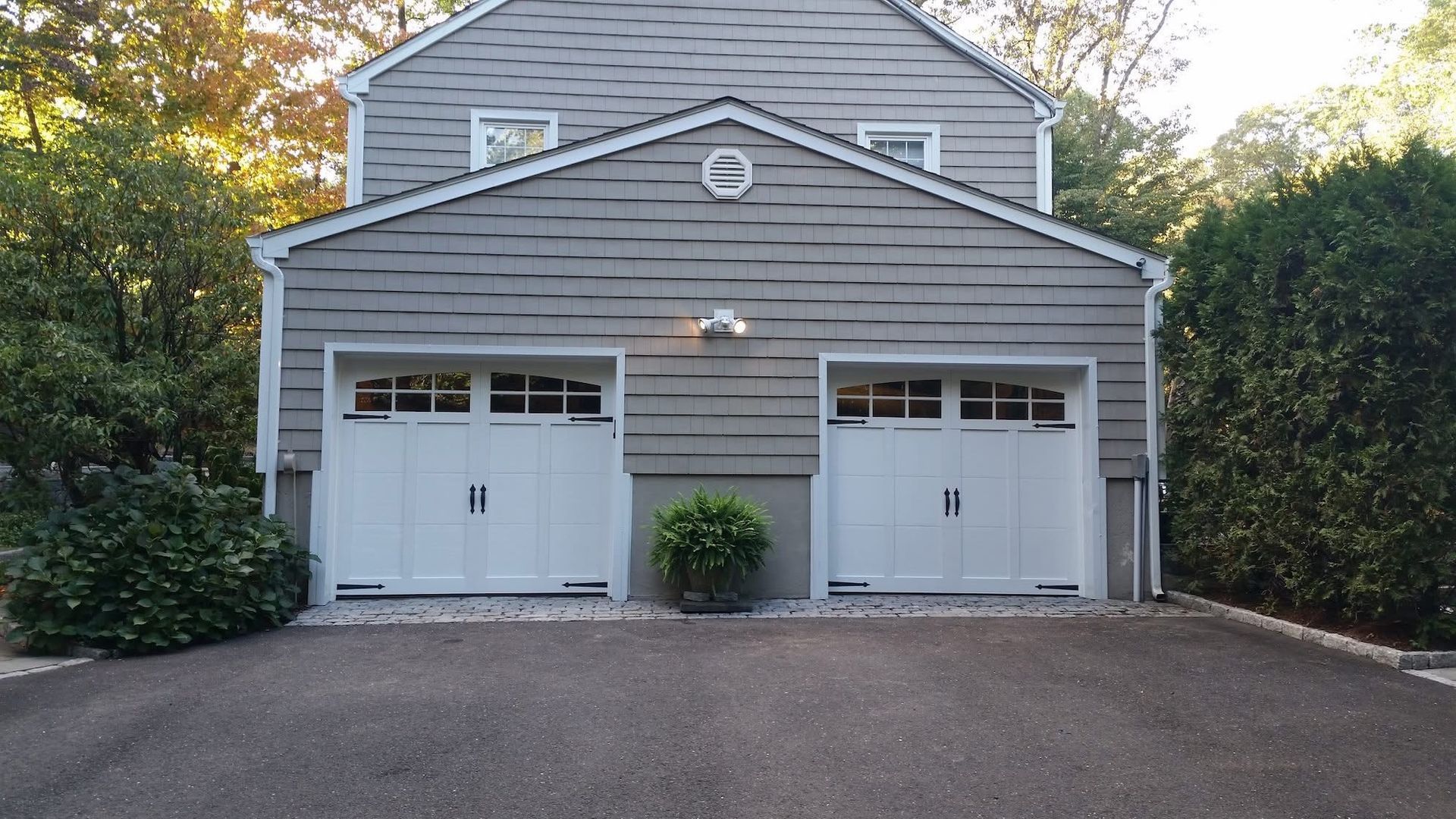 Gray two-car garage with white doors and a driveway, framed by trees and bushes.