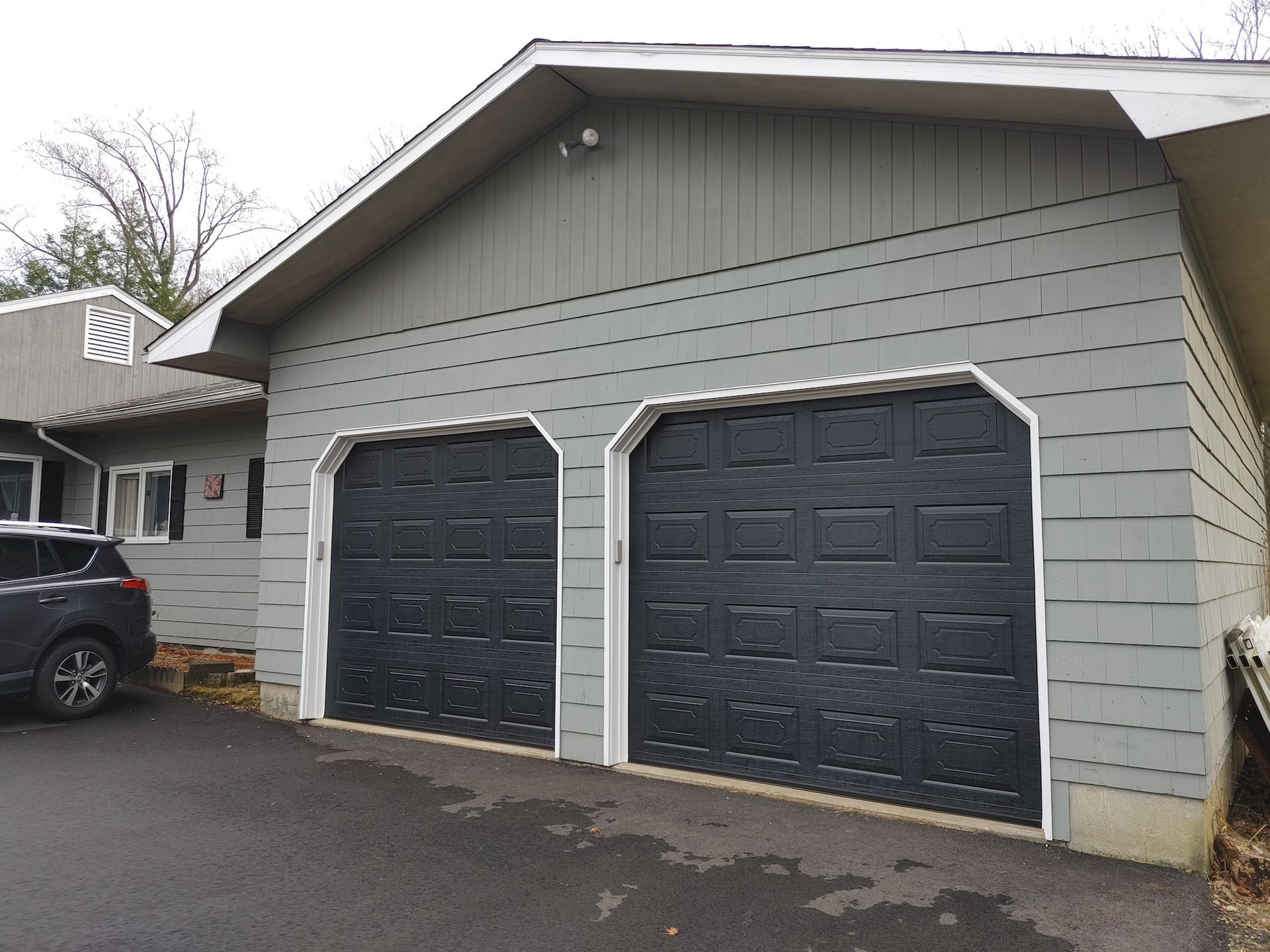 Detached double garage with gray siding and two dark doors beside a house driveway
