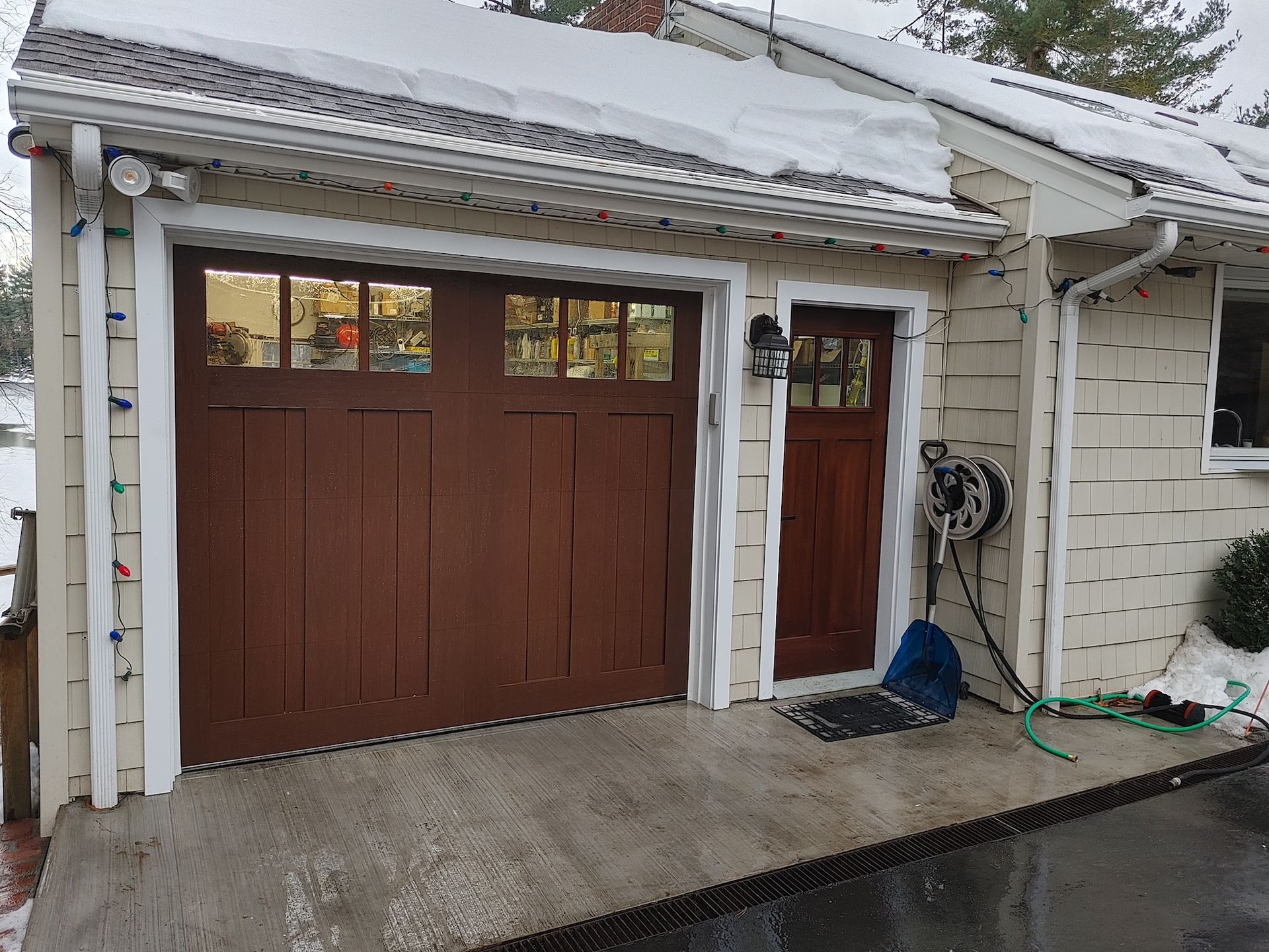 Brown garage with side door under a snowy roof, wet driveway, and hose by the white house wall