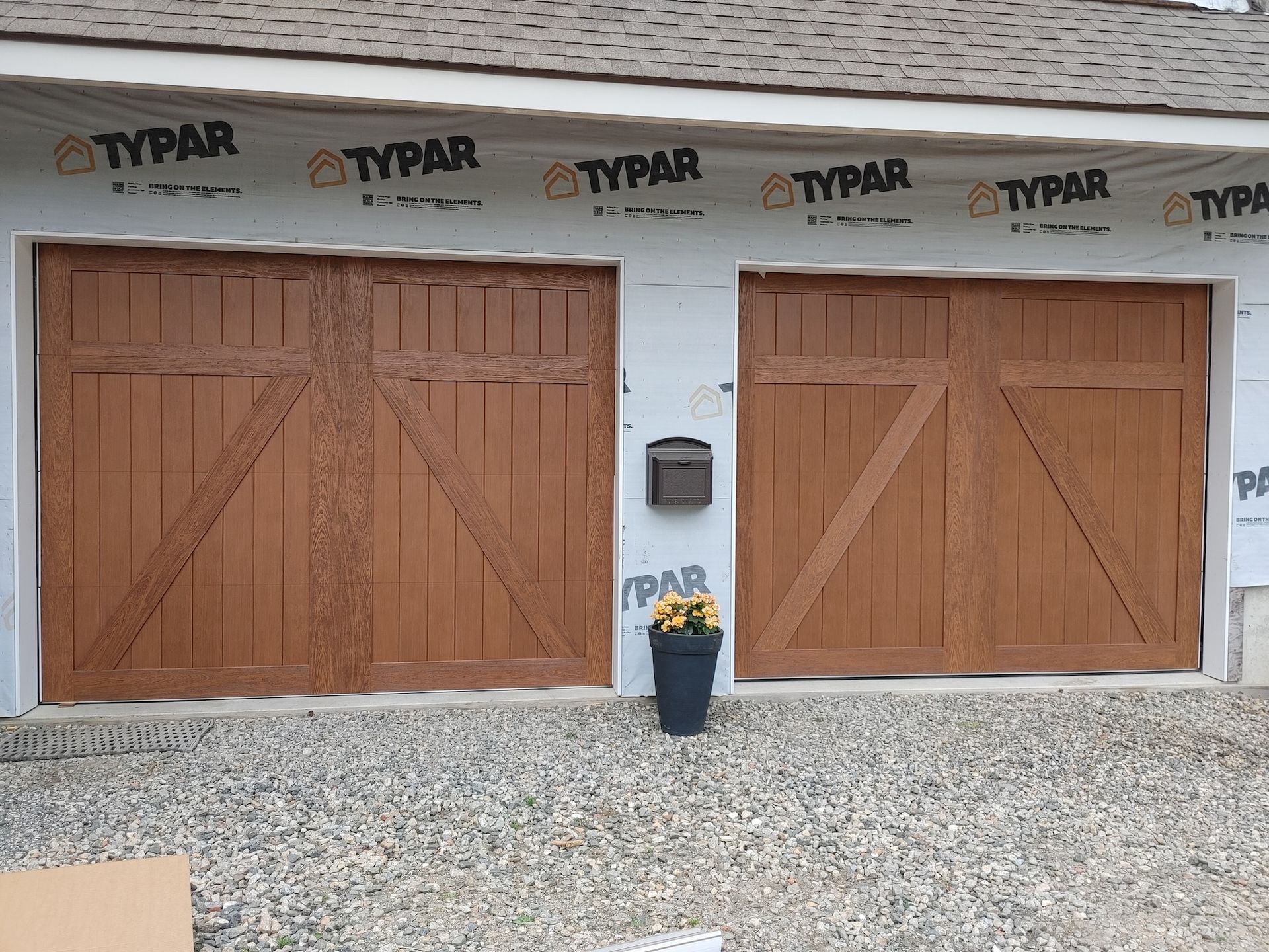 Two wooden garage doors on a house under construction, with Typar wrap and a gravel driveway.