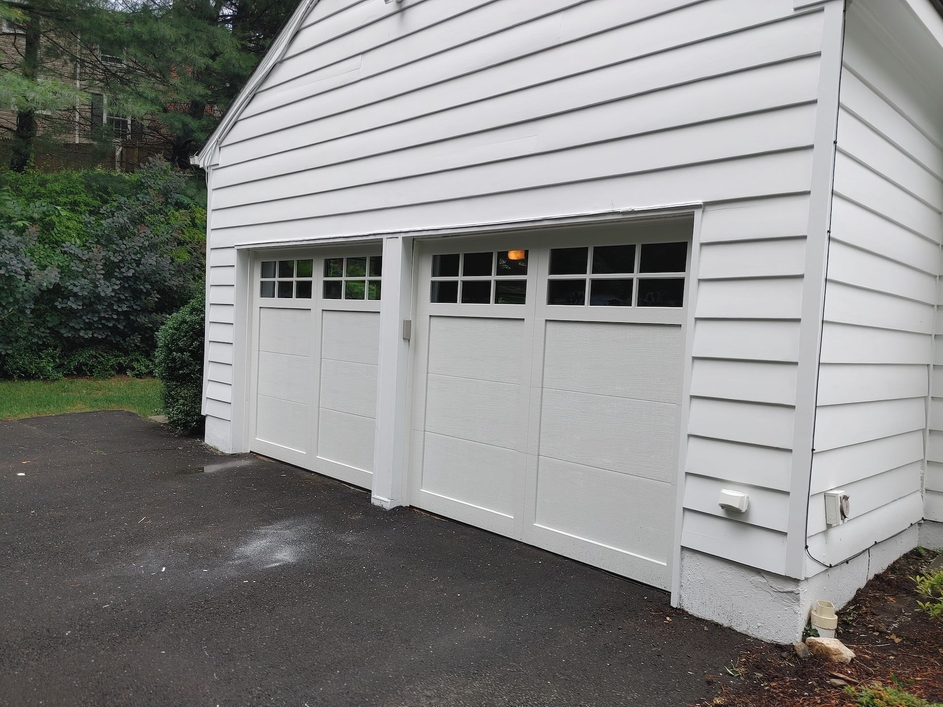White double garage doors on a house exterior beside a wet driveway and trees