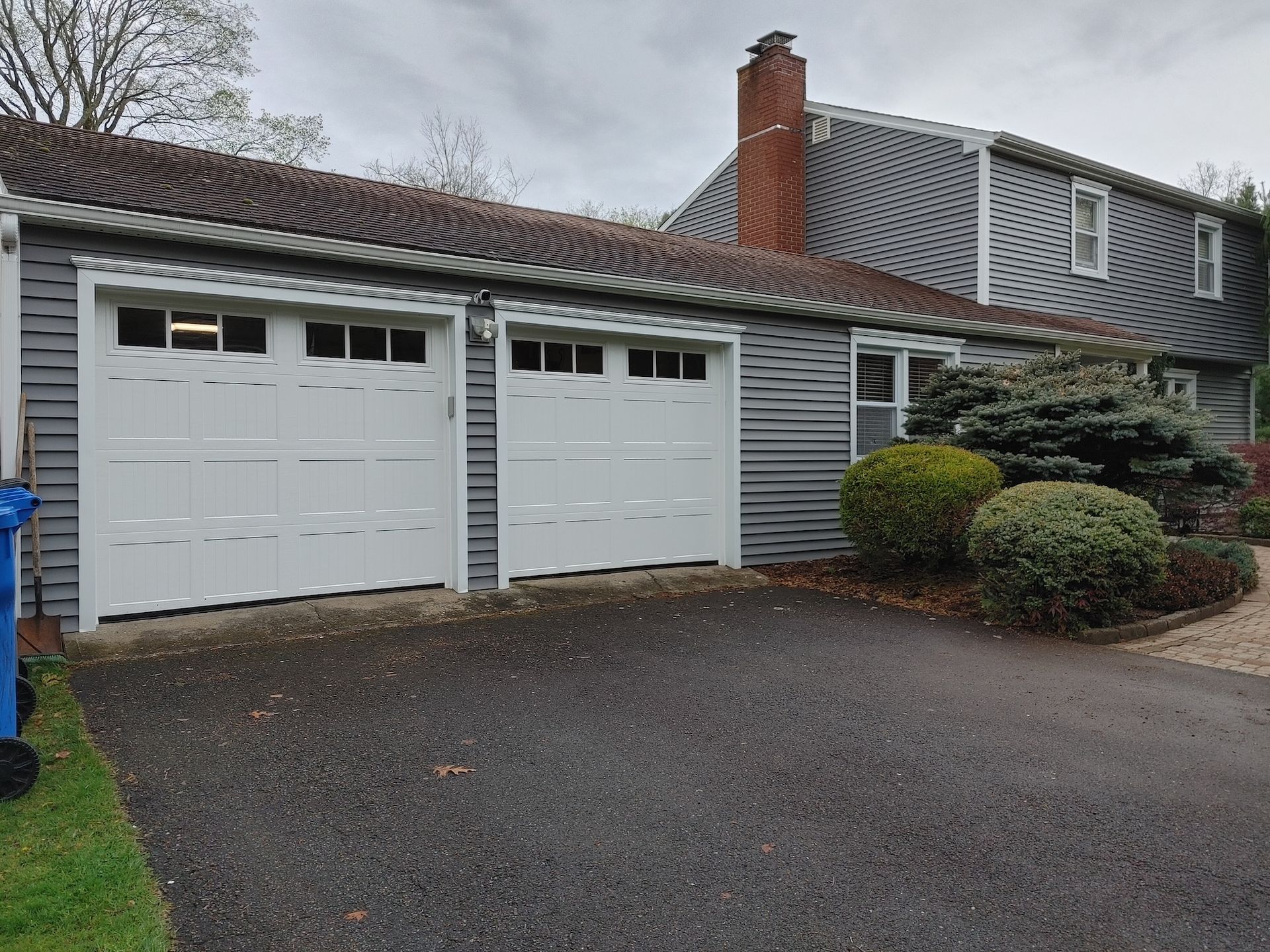 Suburban house with two white garage doors, gray siding, and a driveway under an overcast sky