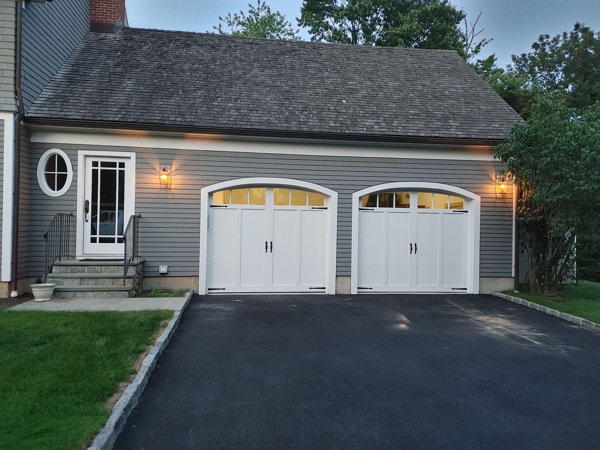 Gray house with two white garage doors, lit porch lights, and a dark driveway at dusk