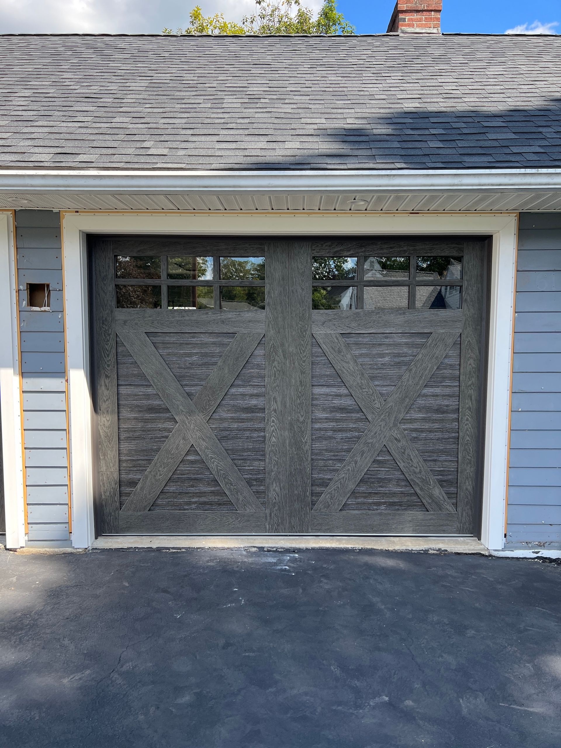 Gray garage door with cross-braced panels on a light blue house, viewed from the driveway
