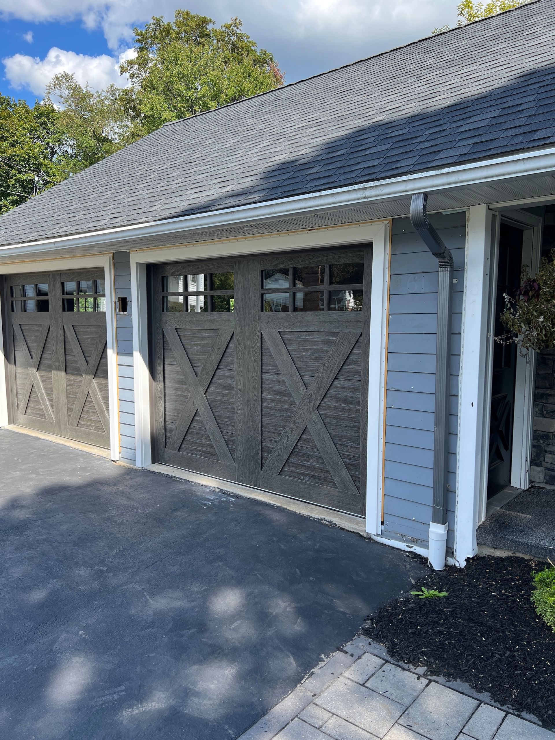 Detached garage with gray shingle roof, white siding, and dark cross-braced wooden doors on a sunny day