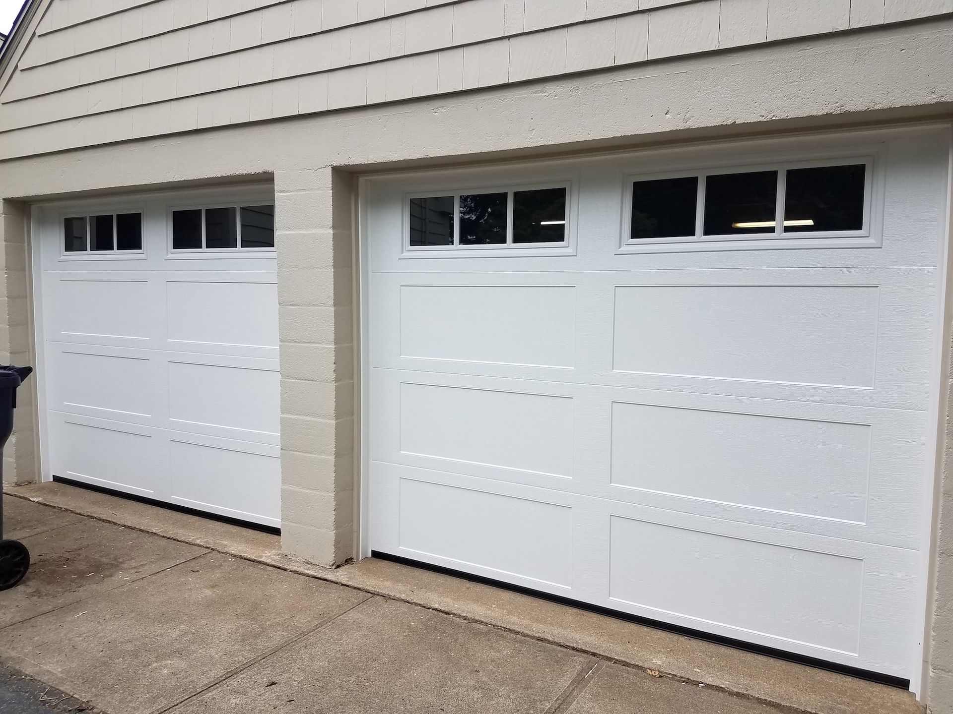 Two white garage doors with small windows in a beige-sided driveway.