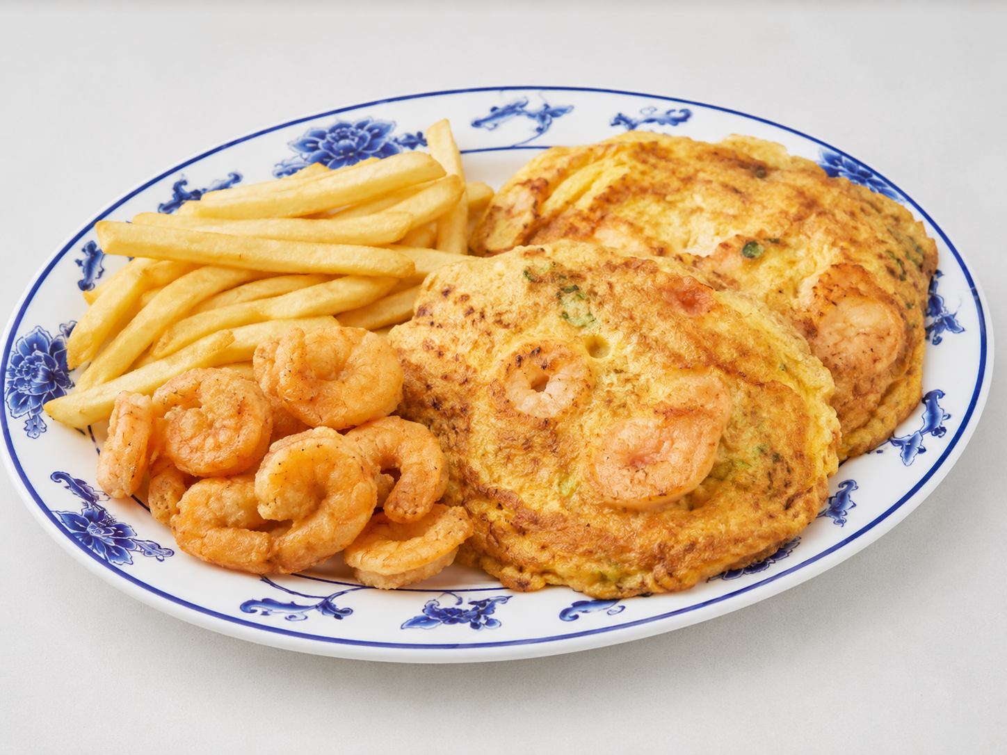Plate of fried shrimp, fries, and two omelets on a white dish with blue floral trim