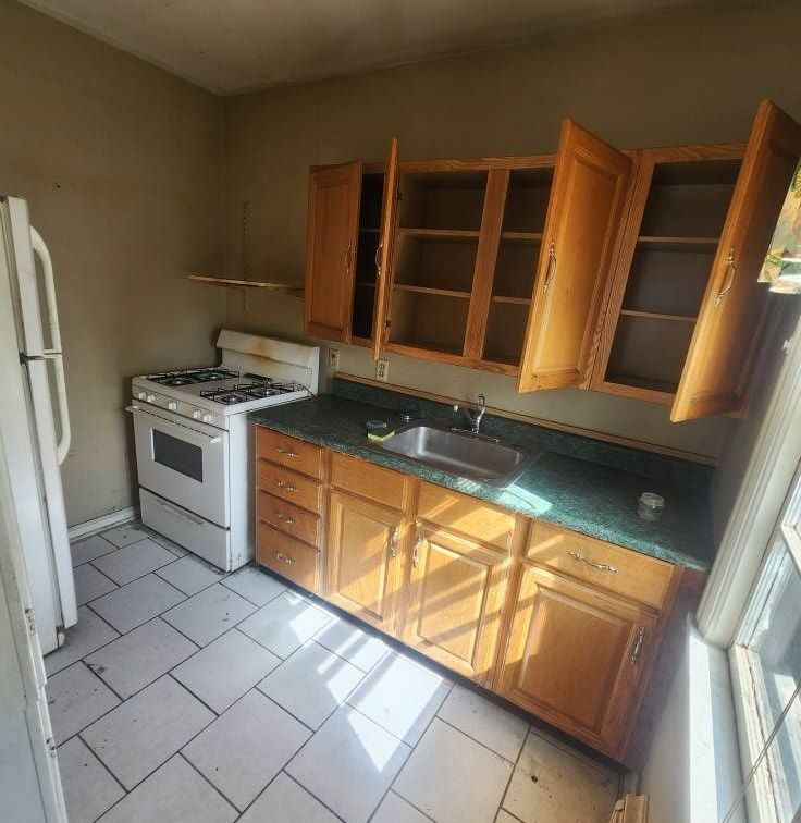 Small kitchen with white stove, wooden cabinets, green countertop, sink, and tiled floor in sunlight