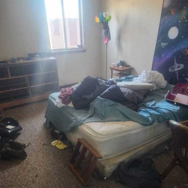 Messy bedroom with an unmade bed, clothes piled on it, and a dresser by a window