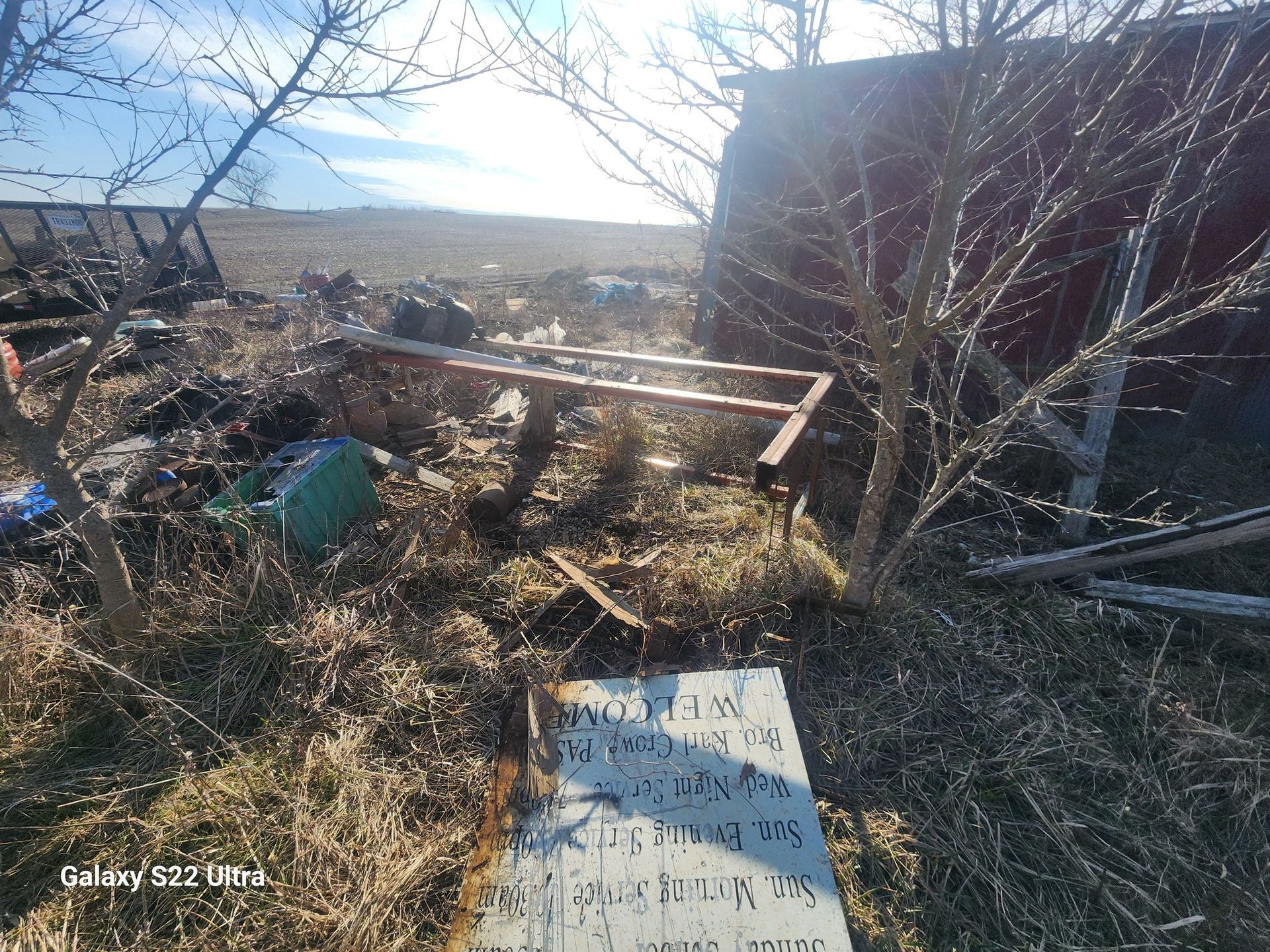 Overgrown yard with debris, a rusted frame, and a weathered sign near a building.