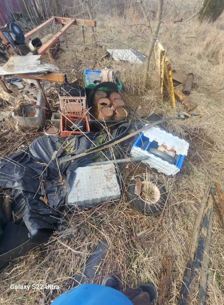 Scattered scrap metal, tires, and debris in a dry, overgrown area near a yellow pole.