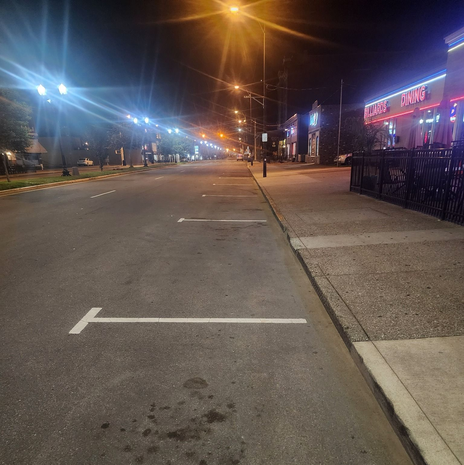 Empty city street at night with streetlights, storefronts, and marked parking spaces.
