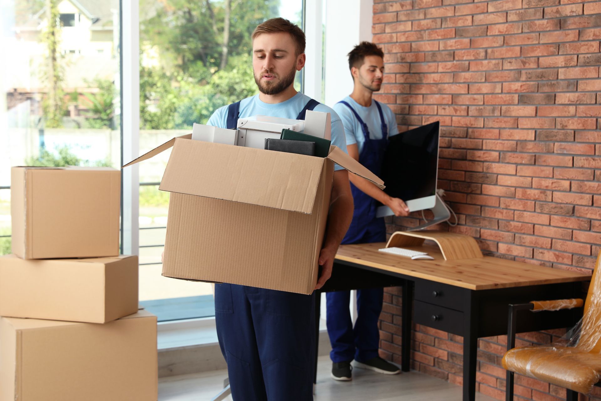 Two movers carrying boxes in a bright office near a desk and computer.