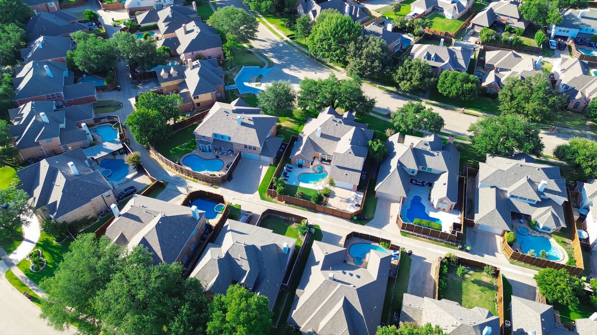 Aerial view of a suburban neighborhood with houses, driveways, trees, and backyard pools.