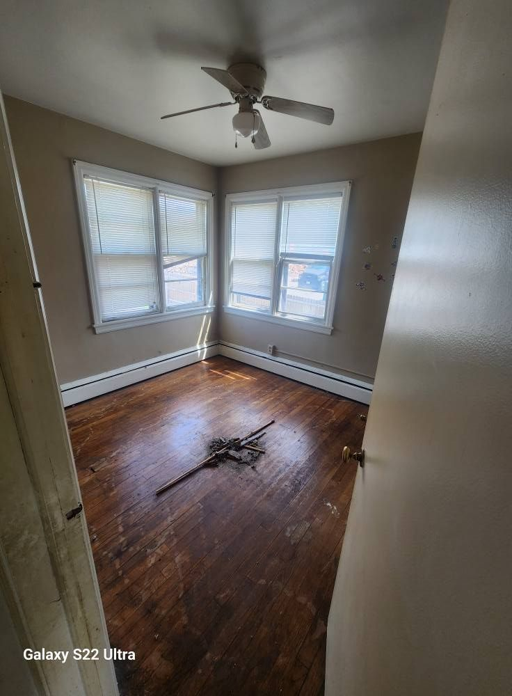 Empty corner bedroom with hardwood floors, beige walls, two windows, and a ceiling fan.