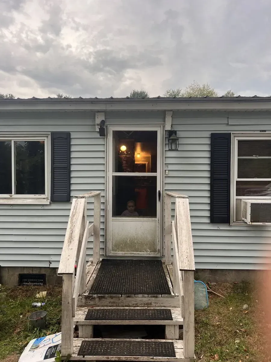 A light blue mobile home exterior with a storm door, wooden porch steps, dark shutters, and a window air conditioner.