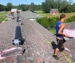 A worker in a blue tank top carries roofing materials on a shingled roof while others work nearby in a residential area.
