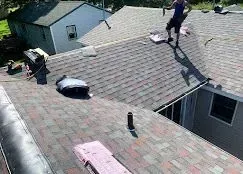 A worker in dark clothing stands on a multi-toned brown shingled roof undergoing repairs, with materials scattered around.