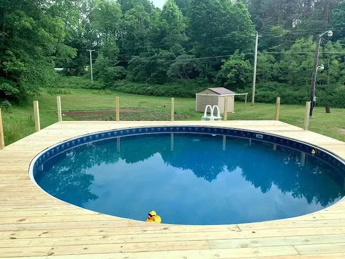 A round, blue above-ground swimming pool surrounded by a newly built wooden deck in a grassy, wooded backyard.