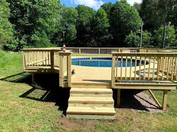 A wooden deck with railings surrounds a round, above-ground swimming pool in a grassy, wooded backyard.