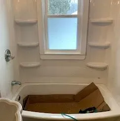 A white bathtub and shower surround with built-in shelves and a window in the center, containing a cardboard liner.
