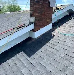 A brick chimney sits on a gray shingled roof with white trim, featuring a wire cable running across the shingles.