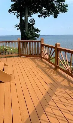 A wooden deck with unique sunburst-style railings overlooking a large, calm body of water under a clear blue sky.