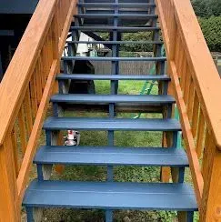 Outdoor wooden stairs with stained railings and blue-painted treads, viewed from above, leading down to a grassy yard.
