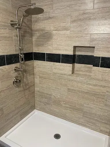 A walk-in shower with tan tiled walls, a black accent stripe, a built-in recessed niche, and a silver shower head.
