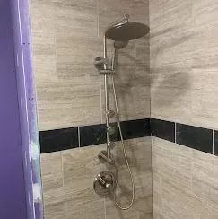 A wall-mounted shower system with a round head and hand shower, set against beige stone tiles with a black accent strip.