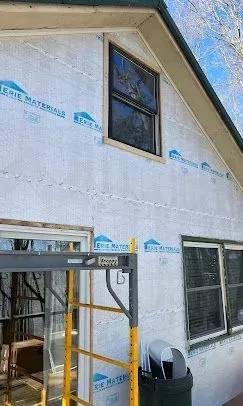 The side of a house under renovation with light gray vapor barrier wrap, two windows, and yellow construction scaffolding.
