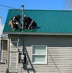 Two people in dark clothing work on the green metal roof of a house, standing near an electrical mast by a ladder.